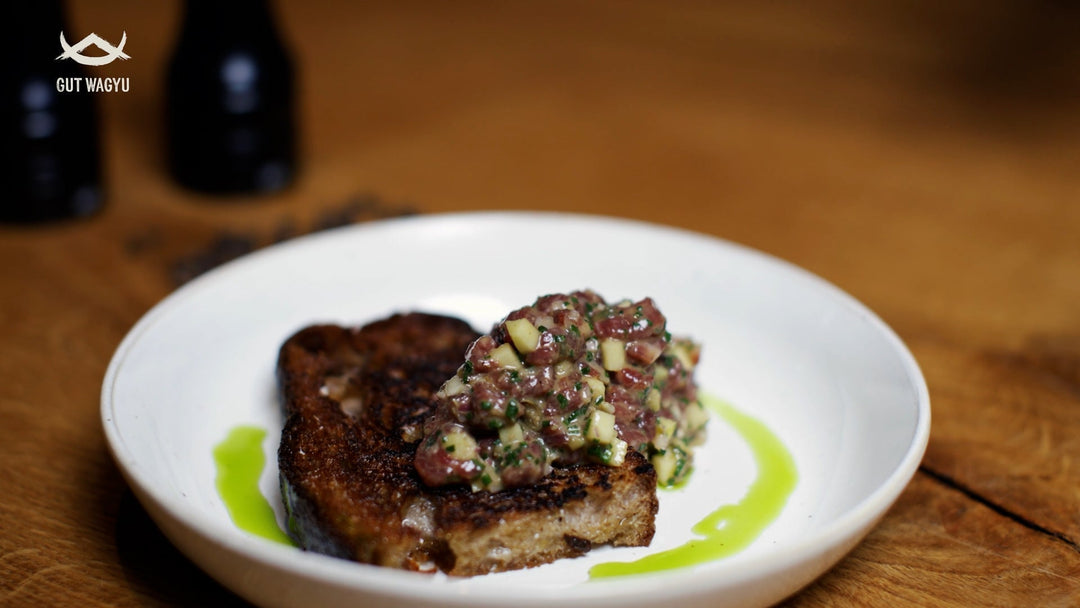 Auf einem weißen Teller liegt Wagyu Tatar vom American Cut auf einem gerösteten Brot, verziert mit Kräuteröl - Daneben Gewürzmühlen auf Holztisch.