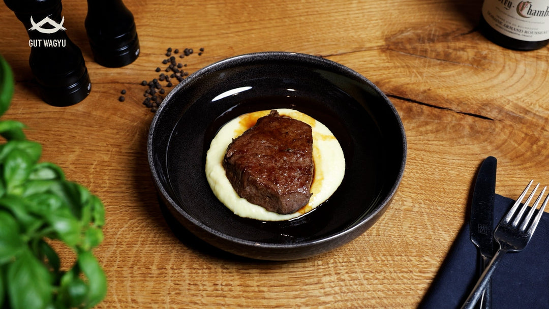 Wagyu Filet auf cremigem Kartoffelpüree daneben Messer und Gabel, Pfefferstreuer und Weinflasche auf Holztisch.