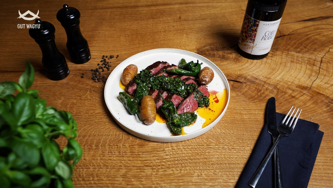 Auf einem weißen Teller liegen Scheiben von gebratenem Wagyu-Rindfleisch, grüne Chillis und kleine Kartoffeln, daneben Pfeffermühlen auf einem Holztisch.
