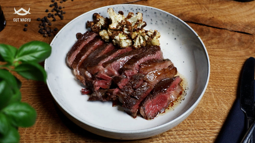 Aufgeschnittenes, rosa gebratenes Wagyu Entrecôte mit geröstetem Blumenkohl auf weißem Teller, daneben Messer und Gabel auf Holztisch.