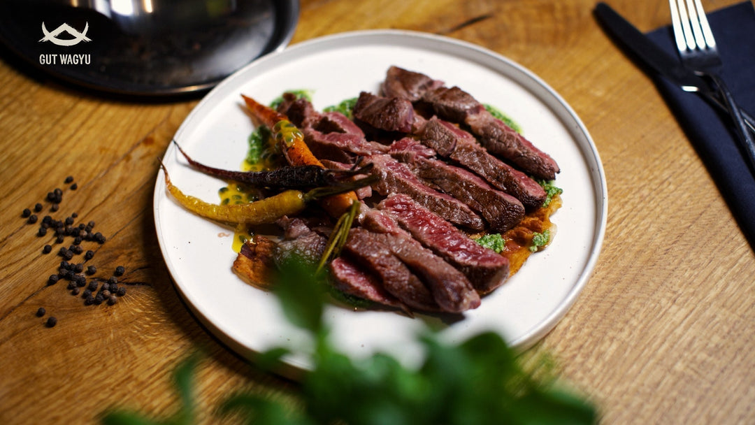 Auf einem weißen Teller liegen mehrere Scheiben rosa gebratenes Prime-Rib-Rindfleisch neben bunten kleinen Karotten, auf einem Holztisch mit Besteck und Pfefferkörnern.