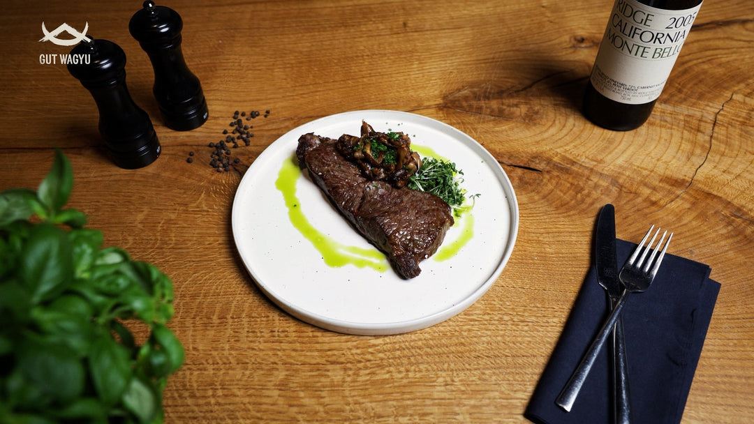 Wagyu Rumpsteak auf weißem Teller mit Pilzen, Kräutern und grüner Sauce, daneben Besteck und Gewürzmühlen auf Holztisch.