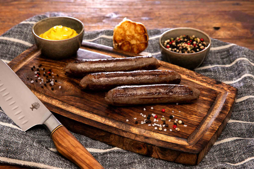 Drei gegrillte Wagyu Bratwürste aus deutschem Wagyu-Rindfleisch von Gut Wagyu, serviert auf einem Holzbrett mit Senf, Knoblauch und Pfeffer.