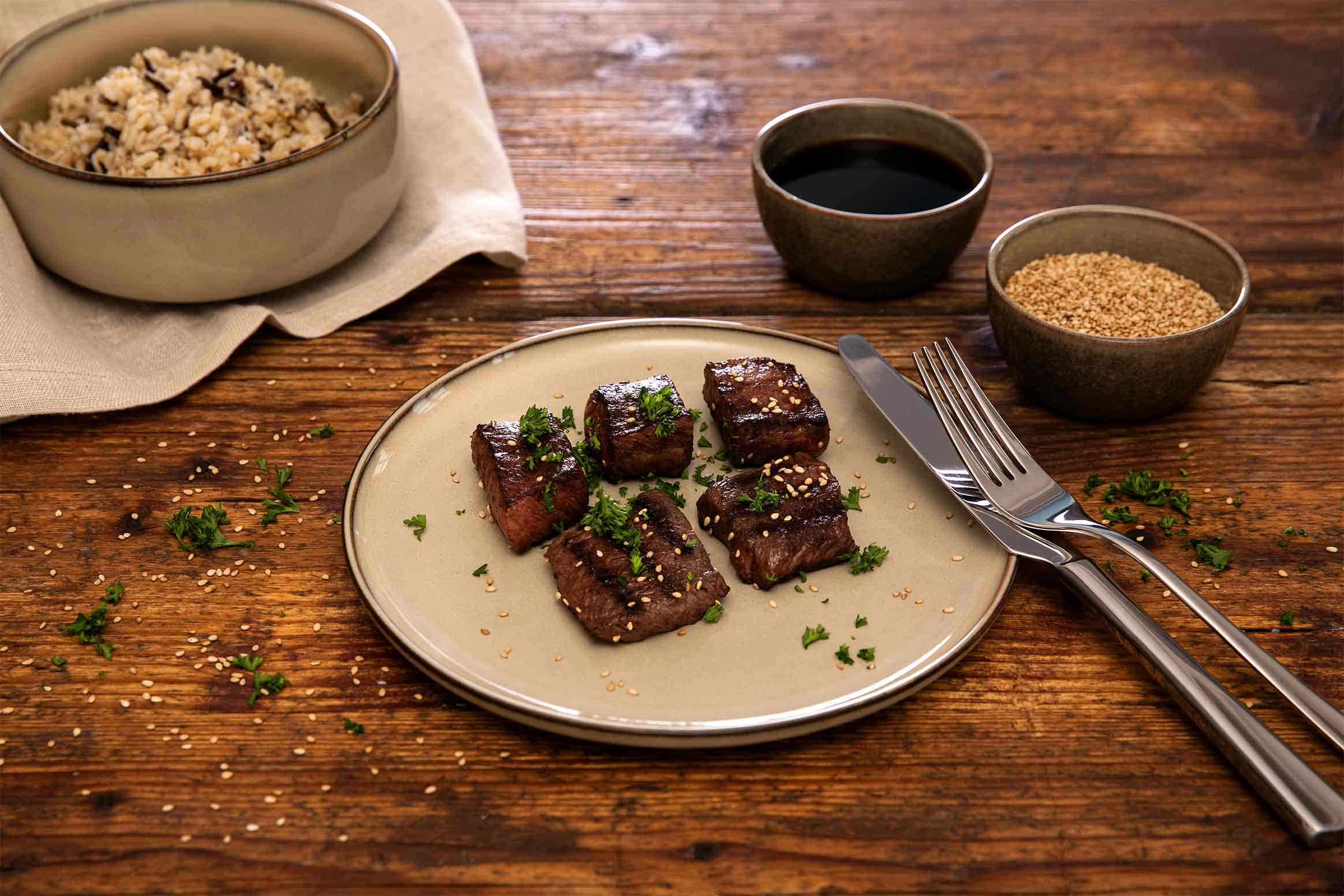 Gegrillte Wagyu Japanese Cut Stücke von Gut Wagyu, mit Sesam und Petersilie auf einem Teller serviert, dazu Reis und dunkle Sauce.