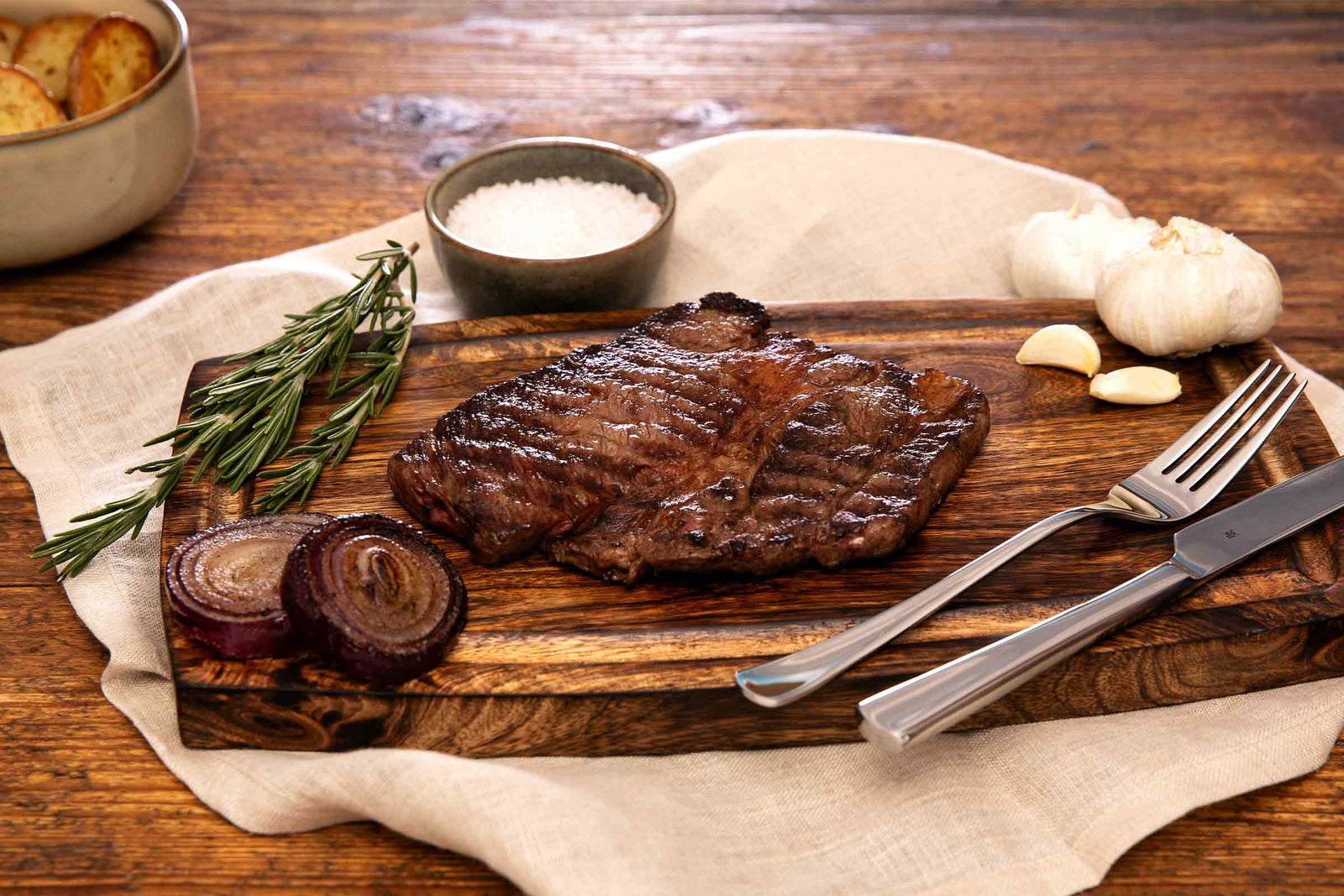 Gegrilltes Prime Rib aus deutschem Wagyu-Rindfleisch von Gut Wagyu, auf einem Holzbrett mit Rosmarin, Knoblauch, roten Zwiebeln und grobem Salz serviert.